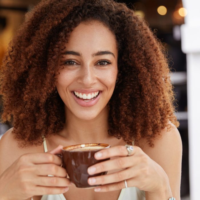 close up portrait of young african american female model has dark healthy skin, white teeth, drinks aromatic espresso, spends leisure time in coffee shop, likes hot beverage. people and lifestyle